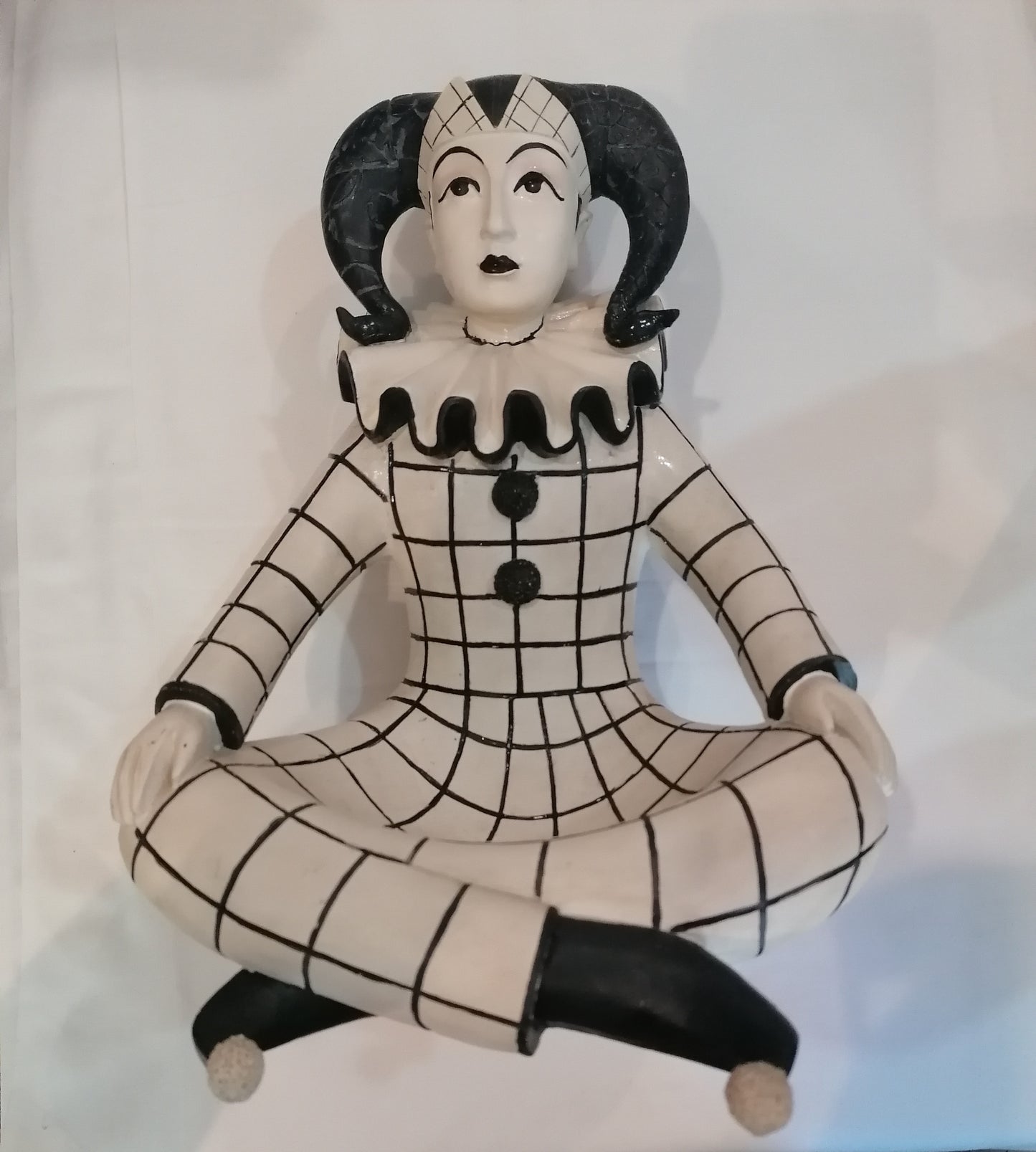 Estátua de porcelana do Arlequim sentado de pernas cruzadas vestido de preto e branco com chapéu clássico da antiga comédia italiana.