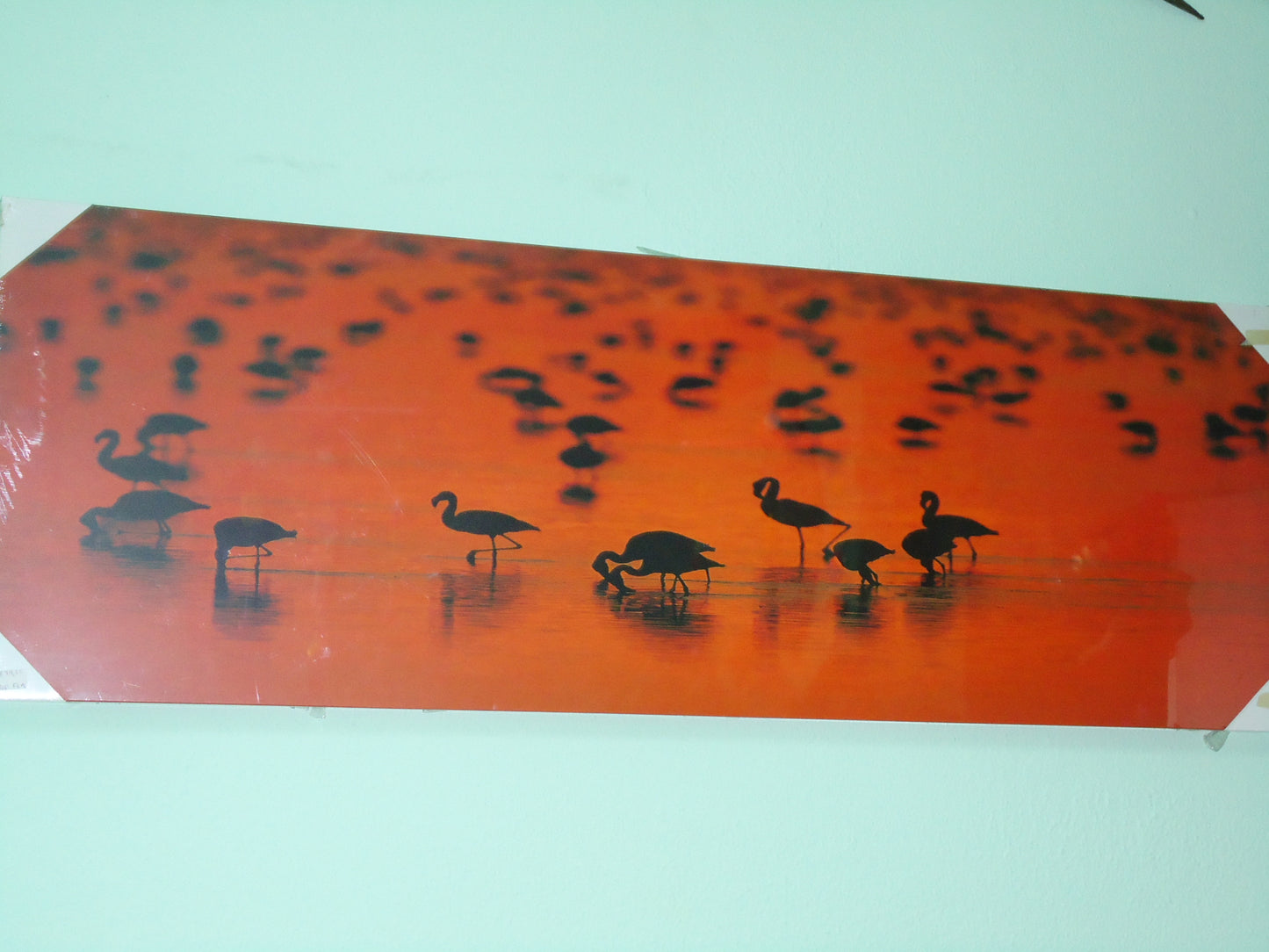 Quadro decorativo com dezenas de silhuetas negras de flamingos num rio cor de laranja vibrante com efeito de contraluz ao pôr do sol.
