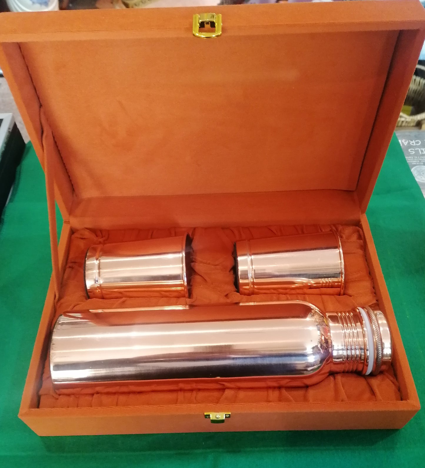 Vista superior de conjunto artesanal da Índia: garrafa de cobre de 950ml e dois copos deitados em berço de caixa  sofisticada de veludo laranja.