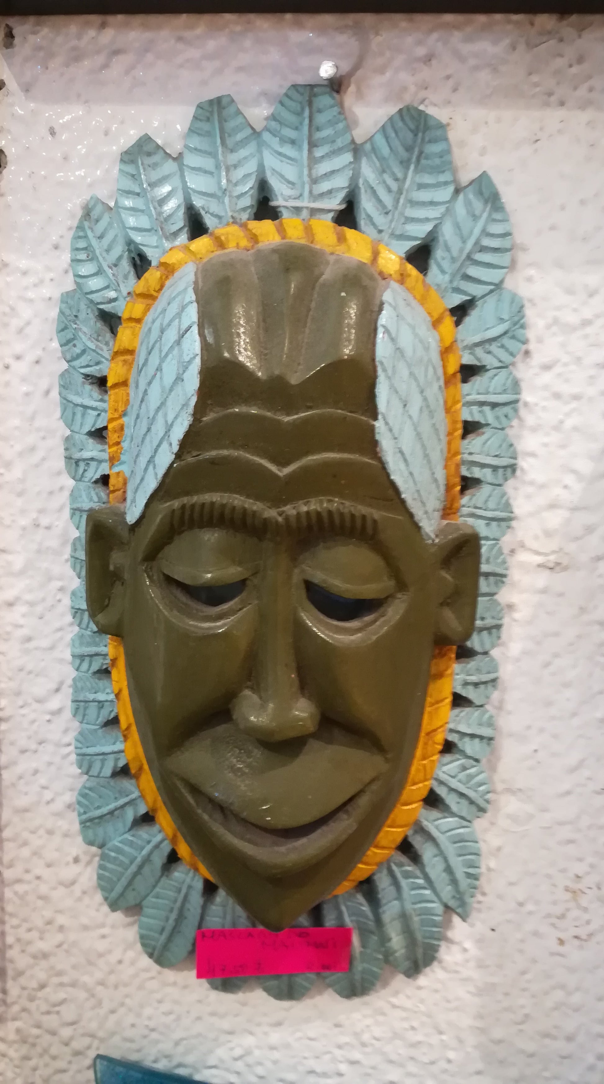 Máscara artesanal africana de parede com o rosto central de um homem cercado por adorno circular de penas decorativas estilo tribal.